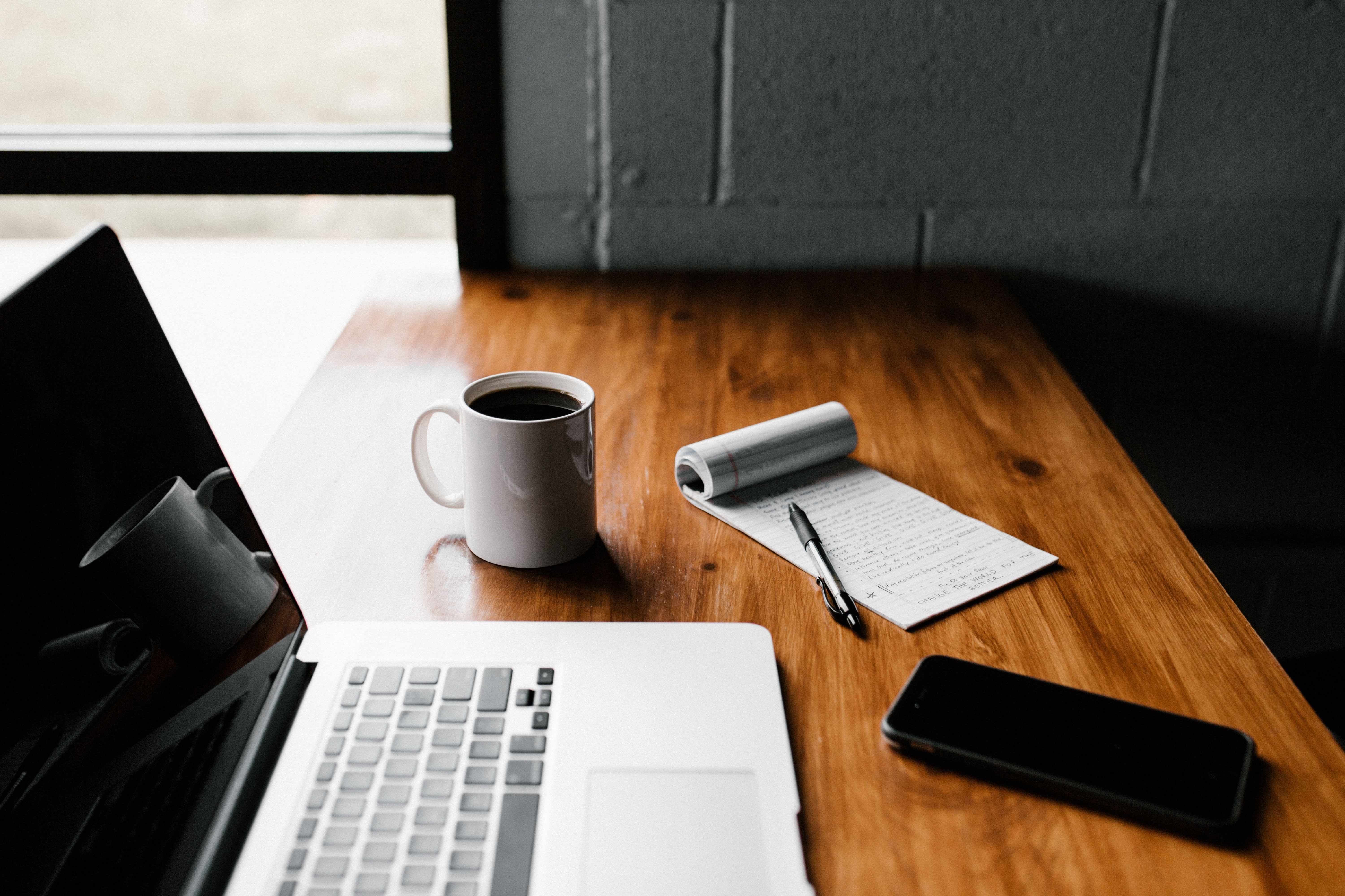 Shows a desk with a laptop, note book, coffee mug and phone